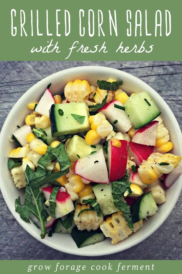 A bowl of grilled corn salad with cucumbers, radishes, and fresh herbs on a wood table.