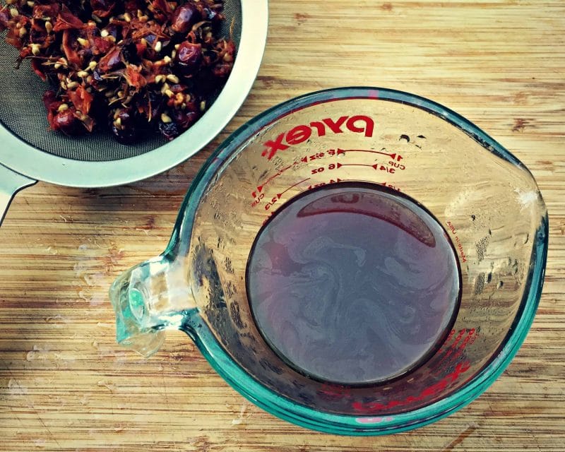 wire mesh strainer with rose hips next to a glass jar of rose hip tea