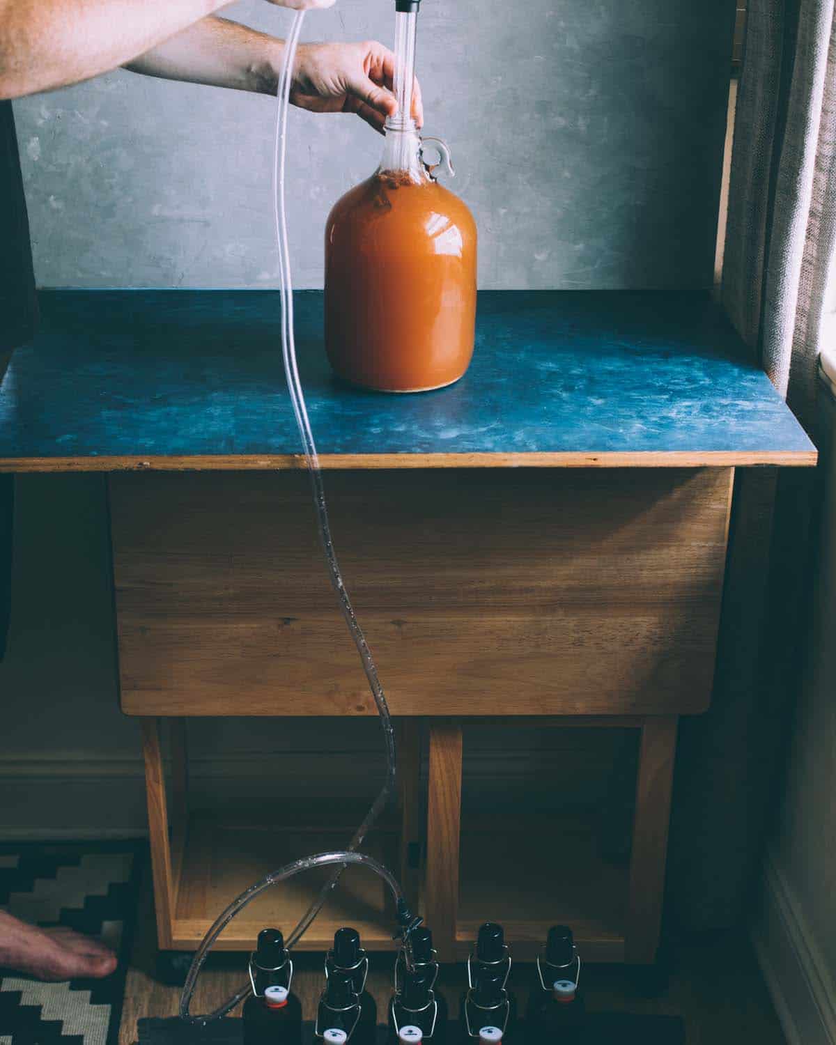 a hand holding the auto siphon in a jug of cider with tubing going down to the bottles on the floor