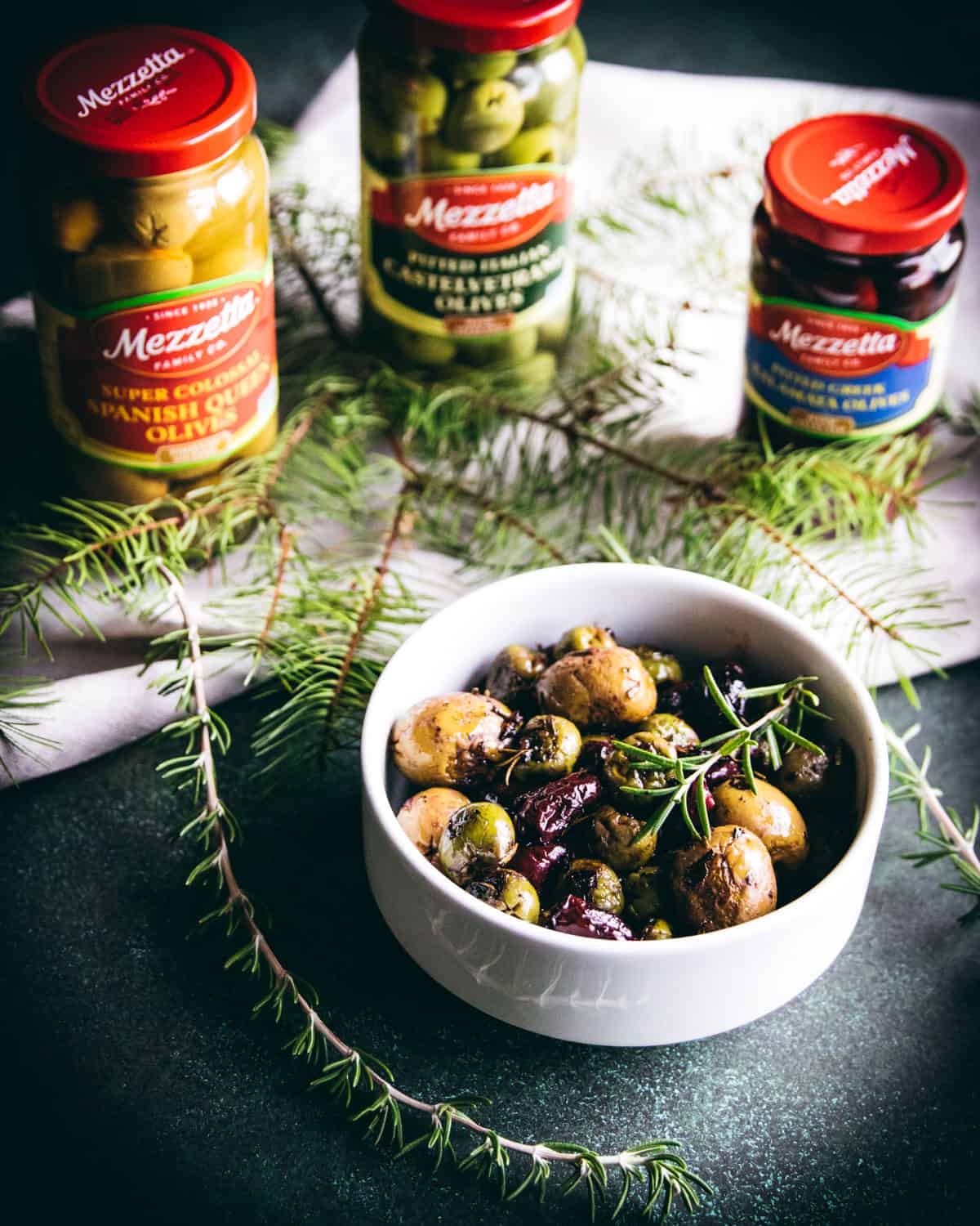 a bowl of roasted olives on a table with some jars of Mezzetta olives