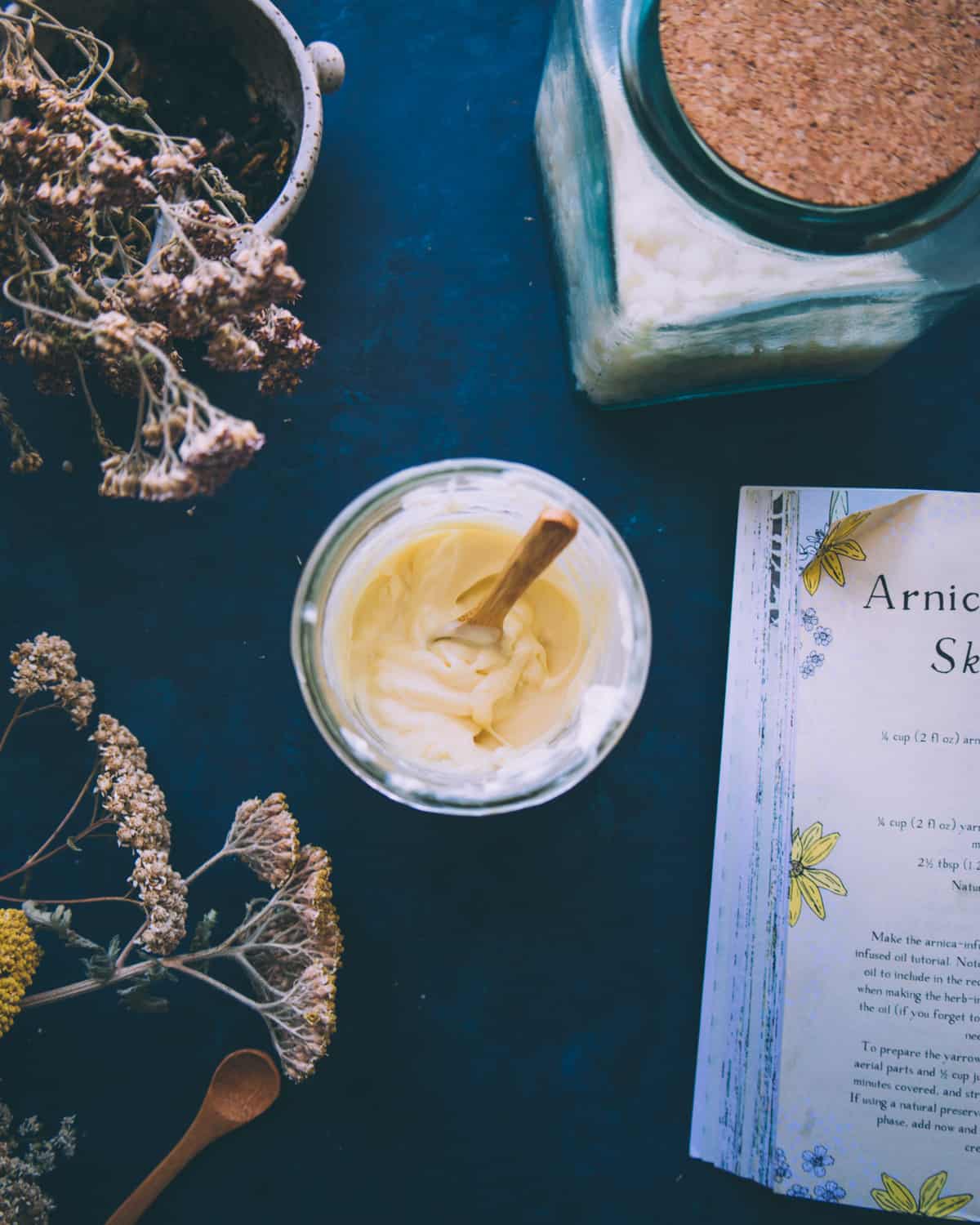 Top view of jar filled with arnica and yarrow cream, with a wooden spoon standing up in it as if it had just been stirred. On a navy blue countertop surrounded by the recipe book and dried yarrow flowers. 