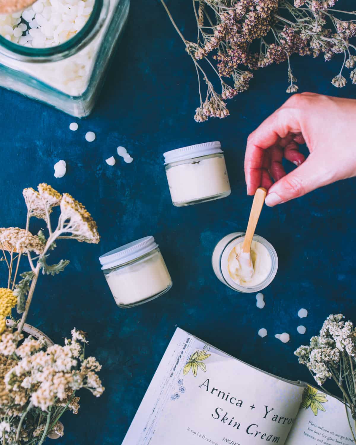 2 jars resting on their sides filled with the cream, and one open top view with a hand stirring the arnica and yarrow cream with a tiny wooden spoon. On a navy blue countertop surrounded by the recipe book and dried yarrow flowers. 