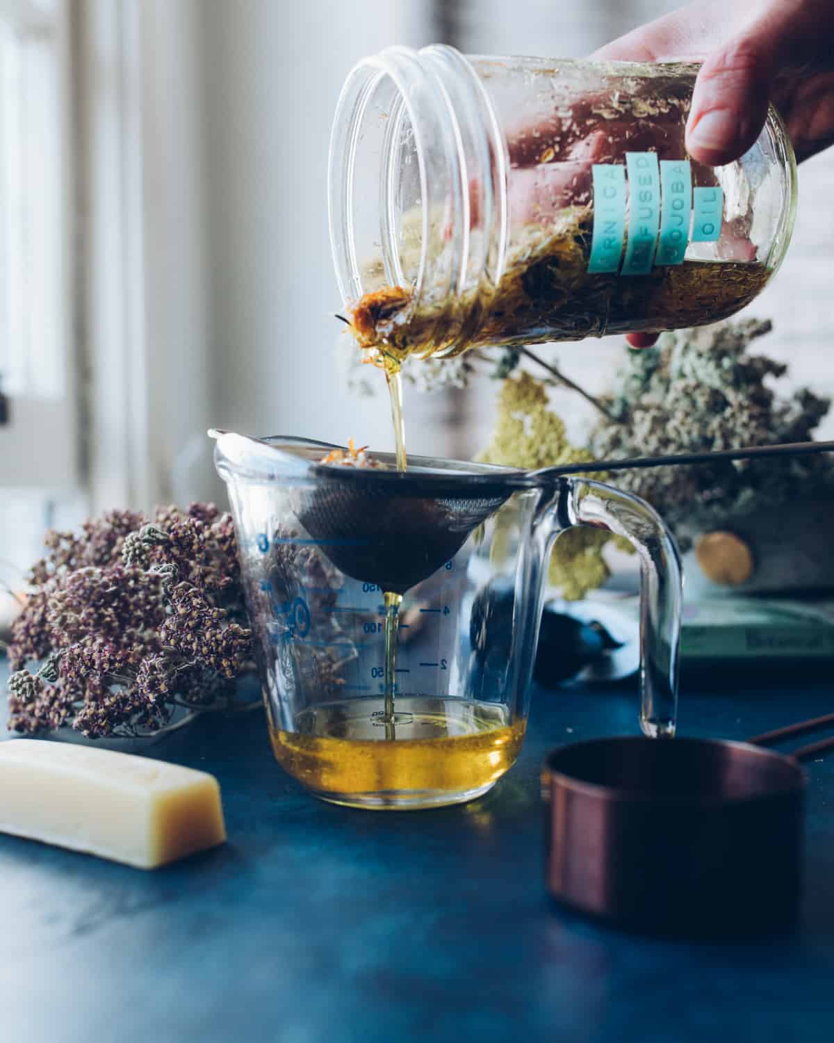 A labeled jar of infused arnica flower oil pouring into a strainer resting in a clear dish, straining the arnica flowers from the oil. On a dark counter, with dried yarrow and a stick of beeswax in the background. 