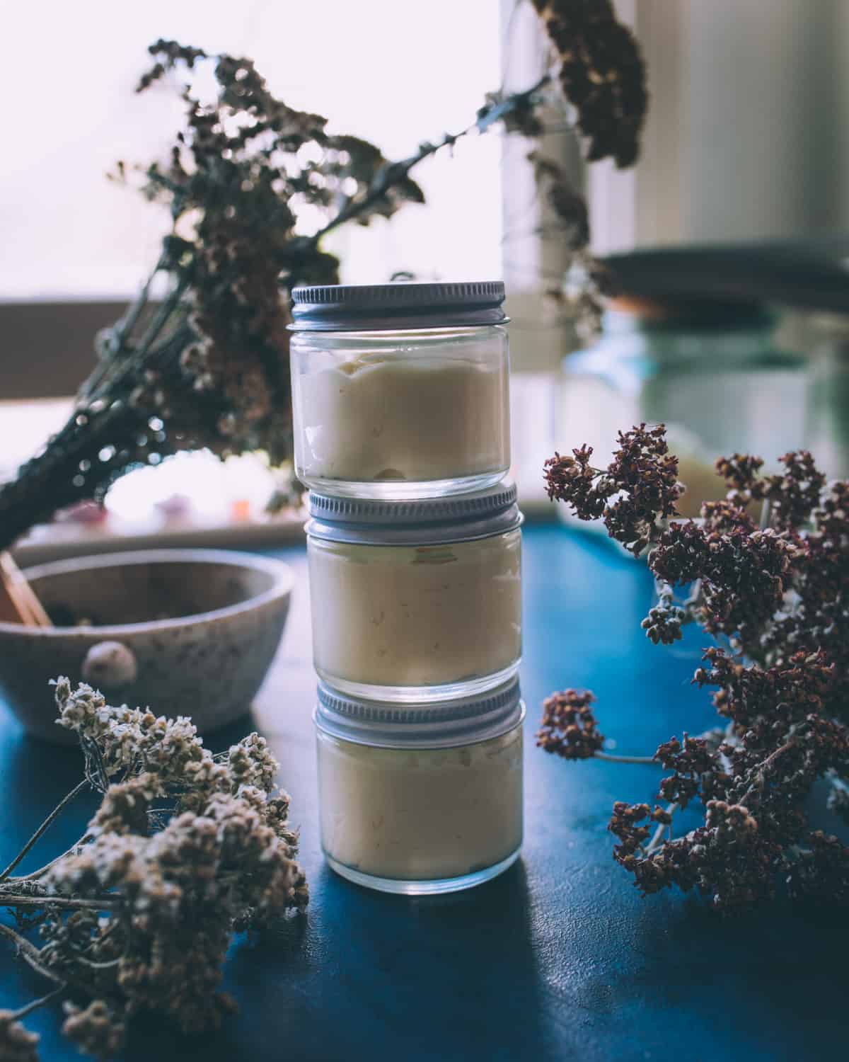 A stack of 3 small jars with lids on, filled with finished arnica and yarrow cream. Sitting on a dark blue counter top with dried yarrow flowers and a mortar and pestle in the background. 