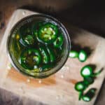 Top view of jar filled with sliced jalapeños and honey, with a wood cutting board and sliced tops of fresh jalapeños in background.